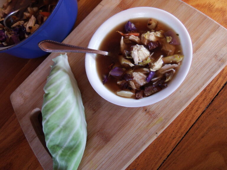 low histamine aip soup with chicken, cabbage, carrots, and broth in white bowl next to a piece of green cabbage on a wood cutting board.